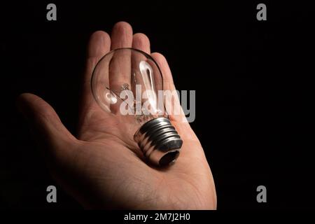 La mano di un uomo tiene una lampadina a incandescenza non funzionante su uno sfondo nero. Mancanza di idee e ispirazione, concetto di burnout Foto Stock