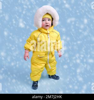 Bambino felice in abiti invernali muta di neve su sfondo blu studio. Un bambino in una tuta gialla calda con cappuccio. Bambino di un anno e cinque mesi Foto Stock
