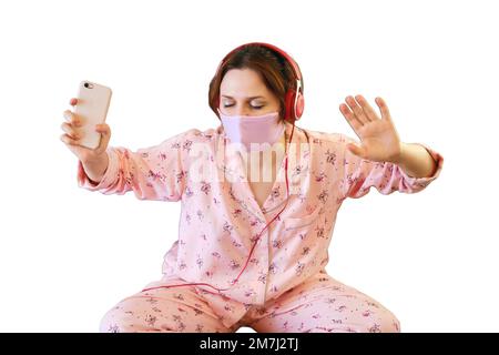 Una donna ascolta musica e balli mentre si siede sul letto durante una malattia con il virus dell'influenza covid, isolato su uno sfondo bianco. Donna in paja Foto Stock