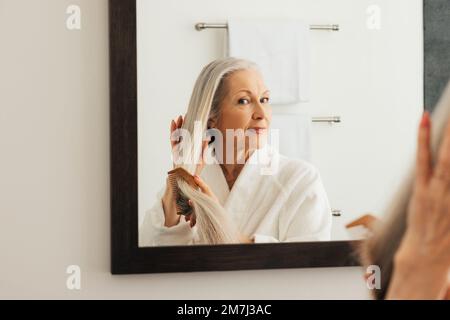 Donna anziana che guarda lo specchio mentre pettina i suoi capelli lunghi in bagno Foto Stock