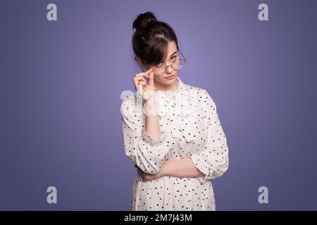 Ritratto di una giovane donna seria e sospetta negli occhiali guardando la macchina fotografica, posando su sfondo viola studio. Foto Stock