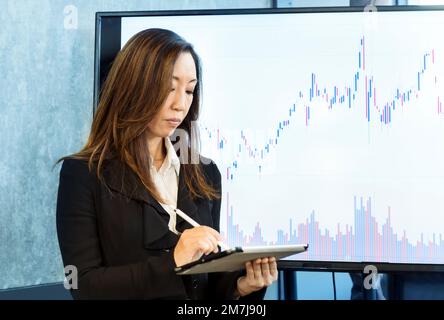 Dirigente etnico concentrato in abito formale vicino a un grande monitor con grafico finanziario mentre si utilizza un tablet in ufficio Foto Stock