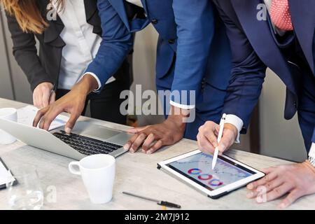 Ritagliate colleghi anonimi in un abbigliamento formale in piedi al tavolo con notebook e tablet mentre lavorate al business plan in un ufficio moderno Foto Stock