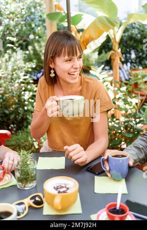 Angolo alto di allegra giovane femmina con tazza di cappuccino ridendo di scherzo e guardando lontano mentre si siede al tavolo durante l'incontro con gli amici in gar Foto Stock
