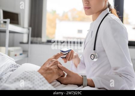 Vista ritagliata del medico che indossa il pulsossimetro sul dito di un anziano in reparto ospedaliero, immagine stock Foto Stock