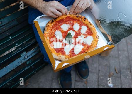 Divertimento rilassato: Uomo che assaggia la pizza napoletana su una panchina da parco da una scatola di cartone. Portate fuori a mangiare in Italia. Foto Stock