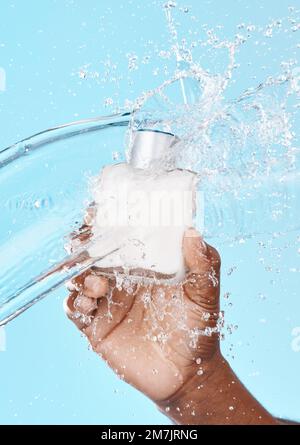 Spruzzi d'acqua, uomo e mano neri con bottiglia per la cura della pelle in studio su sfondo blu. Igiene, pulizia e modello maschile che tiene cosmetici o. Foto Stock
