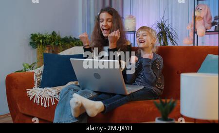 Overjoyed adolescente e piccole sorelle bambini che giocano sul laptop alzano le mani nel trionfo vincitore gesto celebrare successo vincere soldi nel gioco della lotteria. Fratelli bambini o migliori amici in casa sala giochi Foto Stock