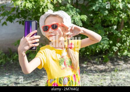 Ritratto di bambina che prende un selfie da smartphone nel parco estivo. Una ragazza allegra in un vestito giallo, cappello bianco e occhiali da sole prende un Foto Stock