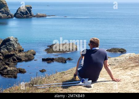 Uomo inriconoscibile disabile seduto sulla roccia con le stampelle accanto, mentre riposa e si gode il bel mare della Bretagna Foto Stock