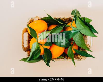 Tangerini e foglie fresche mature in un cesto di vimini su sfondo beige, giacitura piatta. Vista dall'alto sugli agrumi Foto Stock