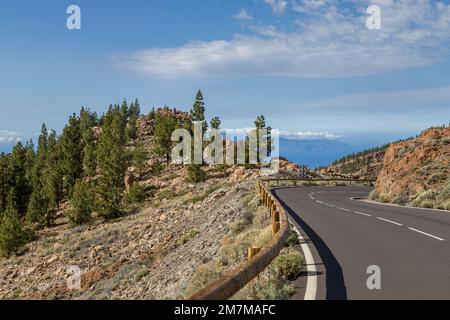 Una collina sul lato della strada con pietre bianche e arancioni e alberi verdi, e l'isola di la Gomera vista in lontananza attraverso la valle, sotto un blu Foto Stock