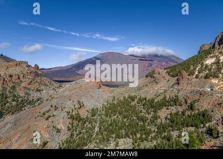 Vulcano di colore viola con il suo picco coperto da una nuvola, visto da una collina sopra il terreno arancione e grigio della valle, con alberi verdi, Foto Stock