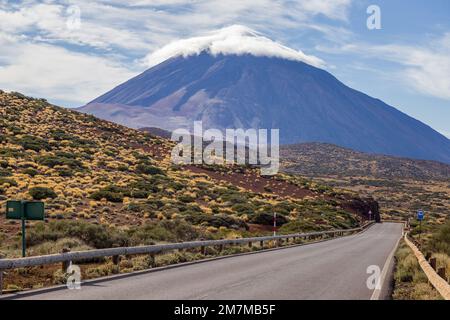 Vista del possente vulcano colorato di blu e viola, con una nuvola bianca che copre la vetta e un paesaggio marrone, giallo e verde coperto di arbusti Foto Stock