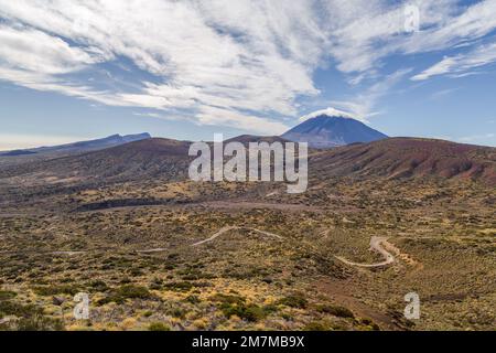 Vulcano colorato blu e viola in lontananza sopra una grande valle coperta di terra marrone e arbusti verdi e gialli, con una strada curva, scoperto Foto Stock