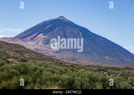 Vulcano come visto da Mirador El Corral del Niño, tinto blu e viola, con campi vicini coperti di terra marrone e arbusti gialli e verdi e bu Foto Stock