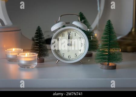 Una sveglia bianca, un puntatore a dodici, un piccolo albero di Natale artificiale verde e candele accese in candelabri su una cassettiera bianca. Copia Foto Stock