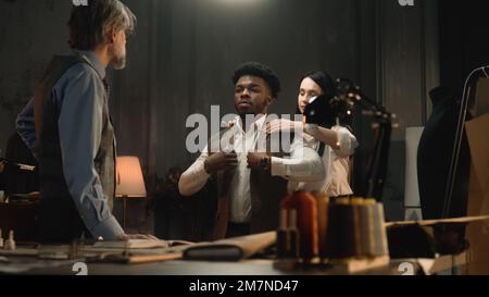 Uomo afroamericano su abito da sposa di affari che si adatta in atelier di lusso o laboratorio di sartoria. Sarti e cliente che discutono il colore e il materiale del giubbotto. Concetto di moda e couturier. Foto Stock