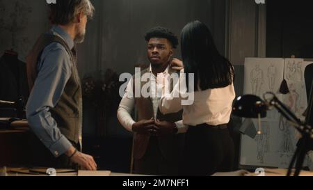 Uomo afroamericano su abito da sposa di affari che si adatta in atelier di lusso o laboratorio di sartoria. Sarti e cliente che discutono il colore e il materiale del giubbotto. Concetto di moda e couturier. Foto Stock