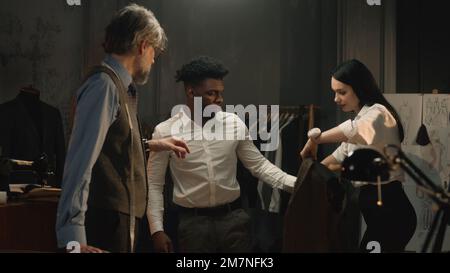 Uomo afroamericano su abito da sposa di affari che si adatta in atelier di lusso o laboratorio di sartoria. Sarti e cliente che discutono il colore e il materiale del giubbotto. Concetto di moda e couturier. Foto Stock