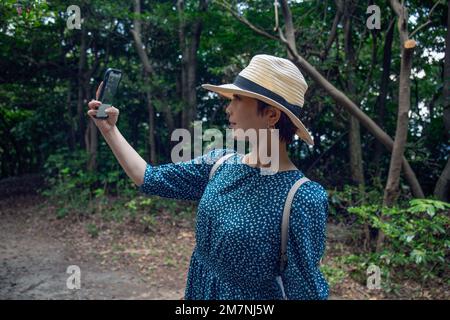 Una donna giapponese matura in un cappello di paglia e vestito blu che prende un selfie con un telefono cellulare. Foto Stock