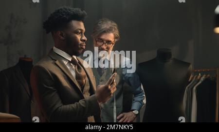 Uomo d'affari afroamericano in studio di sartoria di design di lusso. L'uomo scatta una foto del vestito o registra un video al telefono. Abito da sposa o da lavoro su misura. Moda, artigianato e couturier concept. Foto Stock