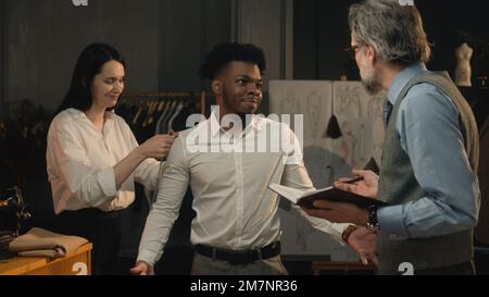 Scatto cinematografico di sartoria che prende le misure dell'uomo afroamericano per la camicia del vestito di affari o di nozze. Lussuoso atelier o studio su misura. Concetto di moda, fatto a mano e couturier. Foto Stock