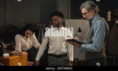 Scatto cinematografico di sartoria che prende le misure dell'uomo afroamericano per la camicia del vestito di affari o di nozze. Lussuoso atelier o studio su misura. Concetto di moda, fatto a mano e couturier. Foto Stock