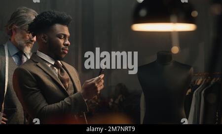 Uomo d'affari afroamericano in studio di sartoria di design di lusso. L'uomo scatta una foto del vestito o registra un video al telefono. Abito da sposa o da lavoro su misura. Moda, artigianato e couturier concept. Foto Stock