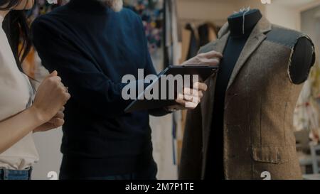 Il fashion designer maschile mostra uno schizzo della futura giacca da vestito a Seamstress sul tablet e discute il processo di sartoria. Manichino in laboratorio atelier. Concetto di moda e artigianato. Primo piano. Foto Stock