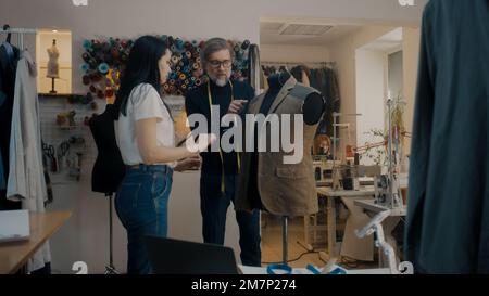 Il designer di abbigliamento maturo con nastro di misurazione sul collo tiene in mano la tavoletta, mostra lo schizzo per personalizzare e discute il vestito. Marinai multietnici che lavorano su macchine da cucire in background. Laboratorio Atelier. Foto Stock