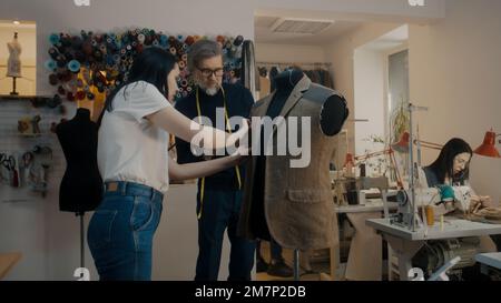 Il designer di abbigliamento maturo con nastro di misurazione sul collo tiene in mano la tavoletta, mostra lo schizzo per personalizzare e discute il vestito. Marinai multietnici che lavorano su macchine da cucire in background. Laboratorio Atelier. Foto Stock