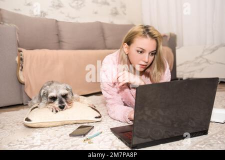 La giovane bionda sta lavorando su un computer portatile, un cane grigio è sdraiato sul tappeto accanto a lei. Foto Stock