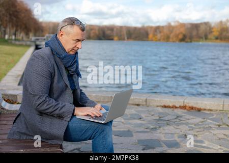 Uomo d'affari dai capelli grigi invecchiato con gli occhiali siedono sulla panca vicino allo stagno dell'acqua che lavora sul laptop. Stile di vita moderno, lavoro remoto. Foto Stock