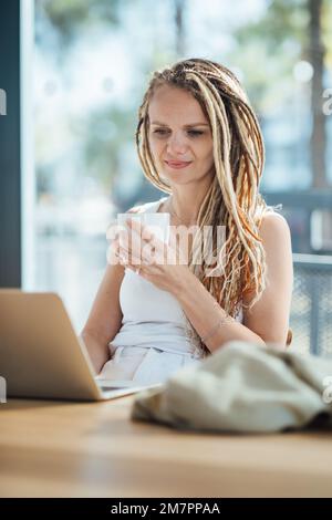 Bella donna sorridente con Dreadlocks che si gode il caffè mentre lavora in caffetteria. Foto Stock