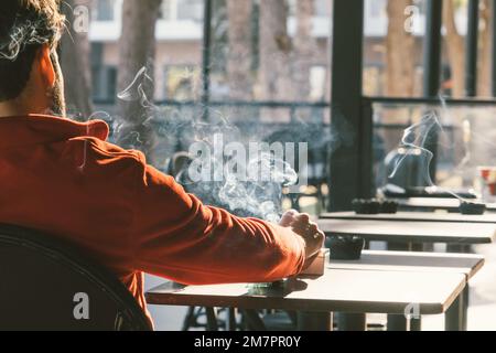 Vista posteriore dell'uomo che fuma una sigaretta di tabacco nel caffè. Stile di vita malsano. Foto Stock