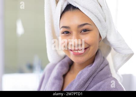 Ritratto di felice donna biraciale con accappatoio e asciugamano sorridente in bagno, con spazio copia Foto Stock