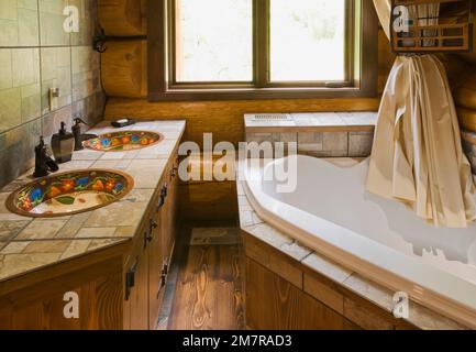 Ripiano in piastrelle di ceramica intarsiato con lavandini in rame verniciato a mano e vasca da bagno nel bagno principale all'interno di una casa in legno di cedro rosso artigianale. Foto Stock