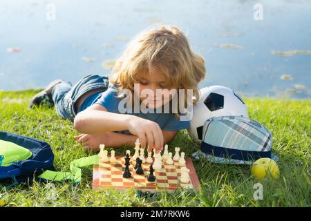 Scuola di scacchi all'aperto. Il bambino pensa o progetta di giocare a scacchi, posando sull'erba nel parco estivo. Ragazzi di scuola intelligenti, intelligenti e intelligenti. Foto Stock