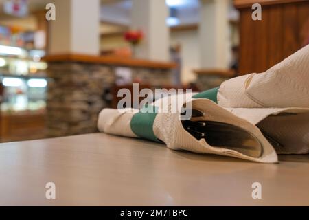 All'interno di un ristorante, le posate si trovano avvolte in un tovagliolo di carta su un tavolo. Gli utensili sono visti da vicino, con la cena sfocata sullo sfondo. Foto Stock