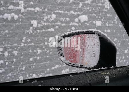 Uno specchio laterale è visto coperto dalla neve bagnata attraverso un finestrino aperto dell'automobile durante una caduta di neve. Foto Stock