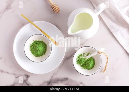 Vista dall'alto di un tavolo in marmo con ciotole di polvere di tè verde opaco organico. una brocca di latte per preparare un latte, un cucchiaio di bambù Foto Stock