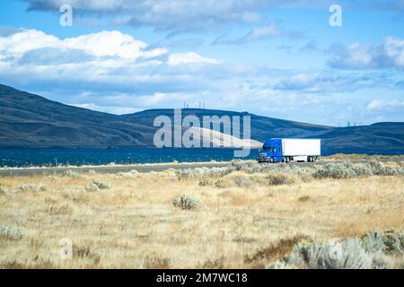 Trattore industriale semi-autocarro grande carro di perforazione blu con cabina alta che trasporta il carico in un semi-rimorchio a secco che guida su strade autostradali divise lungo il fiume wi Foto Stock