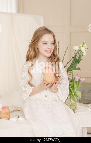 Una ragazza con capelli lunghi in un vestito leggero è seduta al tavolo di Pasqua con torte, fiori primaverili e uova di quaglia. Buona Pasqua Foto Stock