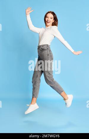 Sentirsi felici. Ritratto di giovane bella ragazza in camicetta bianca e jeans in posa, saltando su sfondo blu studio. Concetto di emozioni, stile di vita Foto Stock