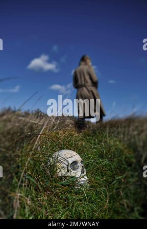 Un cranio umano in parte nascosto in erba lunga con una donna bionda in un lungo cappotto verde in piedi a metà distanza. Stile di copertina del libro di avventure thriller. Foto Stock