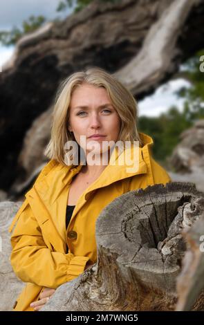 Una giovane donna bionda in un mantello giallo che si erge contro un albero di quercia morto caduto. Foto Stock