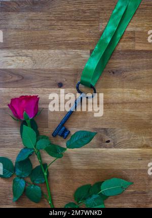 rosa rossa e vecchia chiave con nastro verde adagiato su uno sfondo di legno. ancora vita. Foto Stock