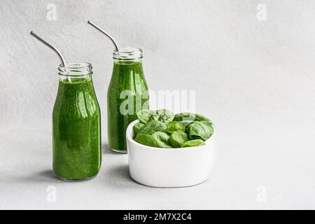 Frullato verde di spinaci in piccole bottiglie di vetro e foglie di spinaci in ciotola su fondo bianco di cemento. Copia spazio per il testo. Concetto di stile di vita sano Foto Stock