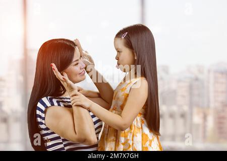 La giovane madre e sua figlia stanno ingannando intorno facendo i volti. La bambina sta mostrando la lingua a sua madre. Sfondo sfocato delle finestre. Foto Stock